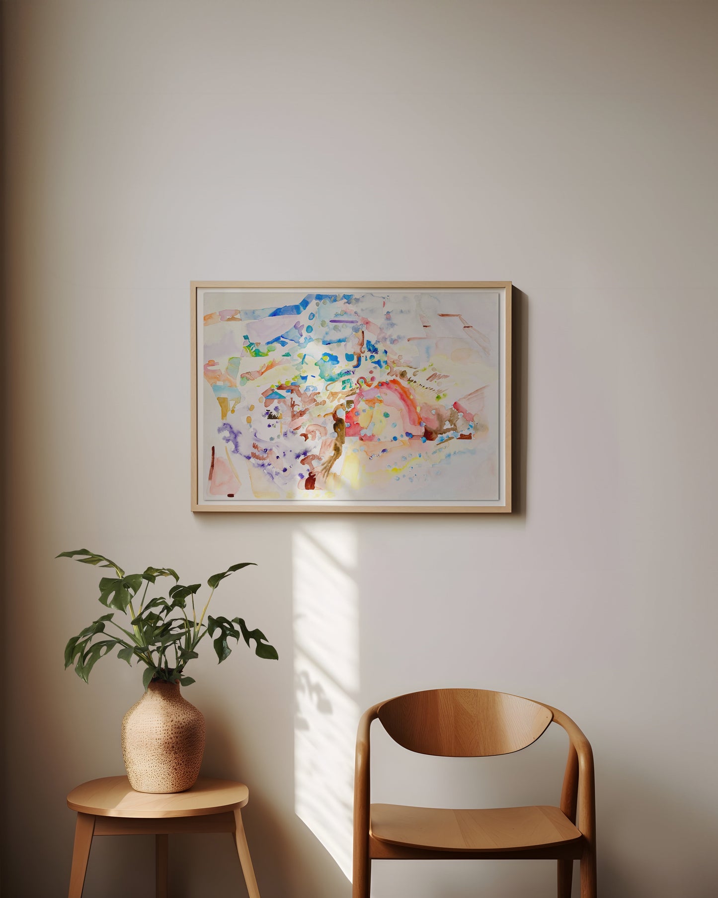 A framed abstract watercolor painting hangs on a beige wall in a styled room setting, featuring a mid-century wooden chair and a small side table with a potted plant.