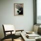 A framed abstract artwork hangs on a light gray wall in a modern living room setting with a wooden chair, a low wooden coffee table, and a window on the right.