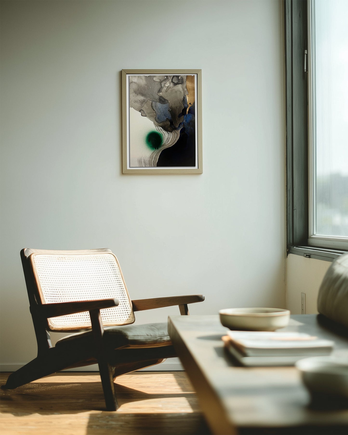 A framed abstract artwork hangs on a light gray wall in a modern living room setting with a wooden chair, a low wooden coffee table, and a window on the right.
