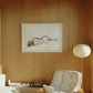 A watercolor painting featuring a nude minimalist figure hanging on a warm wood-paneled wall in a design-focued, sunlit living room with a mid-century white armchair and floor lamp.