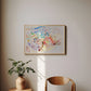 A framed abstract watercolor painting hangs on a beige wall in a styled room setting, featuring a mid-century wooden chair and a small side table with a potted plant.