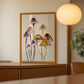 Framed artwork of colorful mushrooms on a wall in a room with wooden furniture and a lamp.
